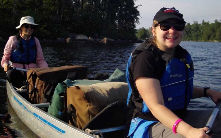 Two young people wearing life jackets smile at the camera as they paddle a canoe.